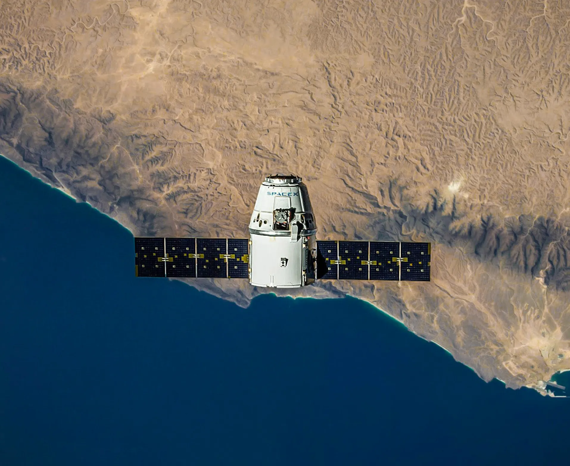 A spacecraft with solar panels orbits above a coastal desert landscape, showing a clear contrast between the land and sea.