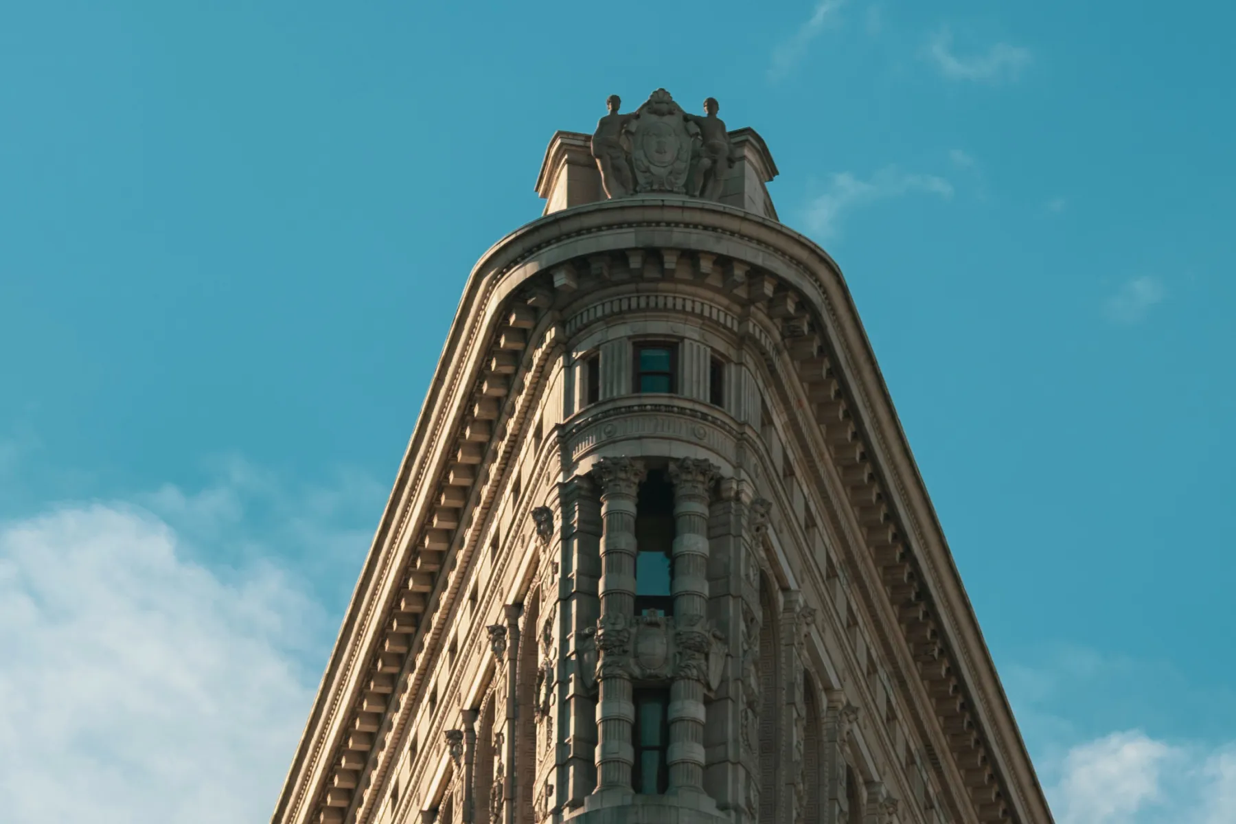 The upper section of a historic building with a flatiron shape, ornate architectural details, and a clear blue sky in the background.