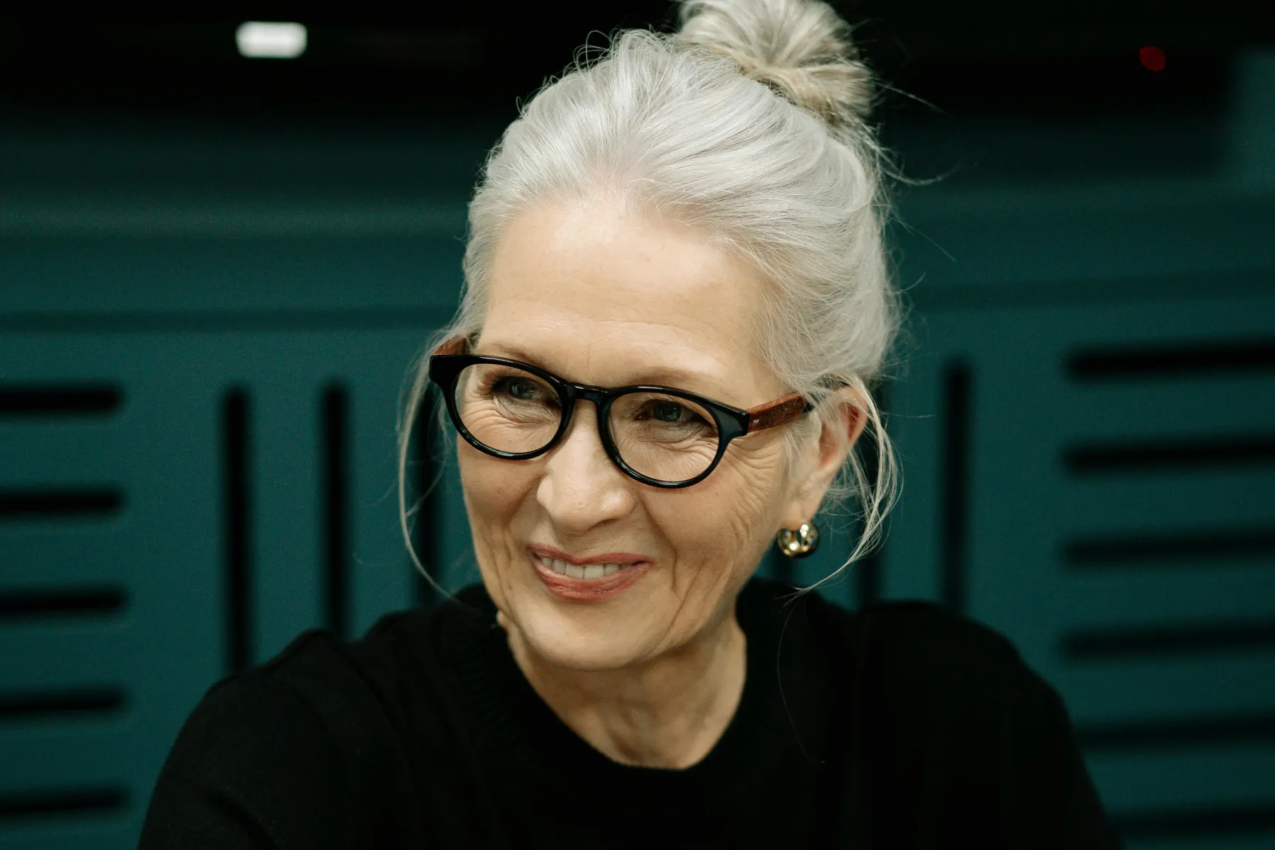 An older woman with grey hair styled in a bun, wearing glasses and pearl earrings, smiles while looking to the side. She is dressed in a black top and is seated in front of a teal background.